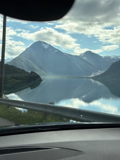 This image captures a serene landscape from the perspective of someone inside a vehicle. The foreground is dominated by the car's dashboard and a section of the windshield, along with a metallic guardrail. Beyond this, a calm, reflective body of water stretches out, mirroring the sky and the majestic mountains that form the backdrop. The mountains are imposing, with slopes covered in lush green vegetation on the lower parts, transitioning to snow-capped peaks higher up. A small village or settlement is visible nestled on the side of one of the mountains. The sky is a dynamic mix of blue patches and scattered white clouds, illuminated by sunlight, suggesting it is daytime. The scene is likely located in or near Halsa, Norway, given the characteristic mountainous fjord landscape. There are no people or animals visible in the frame.