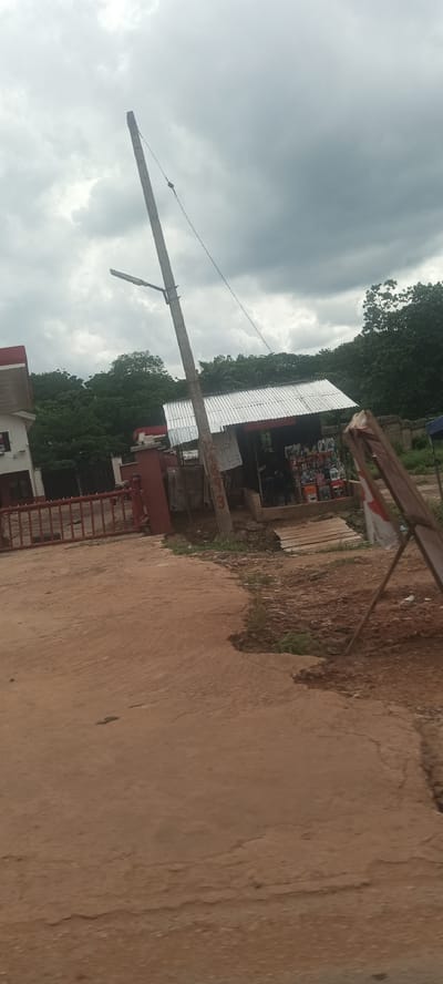 The image depicts a street scene in Ozalla, Nigeria. The foreground is dominated by a dirt road or pathway, appearing worn and uneven.
To the left, a red metal fence and a building with a red roof are partially visible. A leaning utility pole stands near the fence, equipped with a street light.
To the right, a small roadside shop with a corrugated metal roof is present. The shop displays various products, including motor oil containers. An advertisement board is leaning against the shop's exterior.
The background features dense green trees, suggesting a rural or semi-rural setting. Overcast skies fill the upper portion of the image.