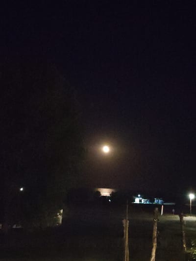 The image depicts a nighttime scene in Limani Litochorou, Greece, characterized by darkness and limited illumination. The sky is predominantly black with a single, bright moon positioned in the upper center of the frame. In the background, several structures with illuminated windows are visible, suggesting a populated area. A streetlamp with a bright light source is situated on the right side of the image, casting a warm glow. The foreground features silhouetted vegetation and wooden posts, possibly part of a fence or boundary. The overall ambiance is one of quiet and solitude under the moonlight.