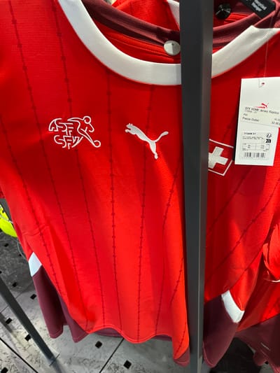A red Puma Switzerland national football team jersey is displayed on a hanger in a retail store. The jersey features a white crew neck collar and white Puma logos on the chest. The Swiss Football Association (SFA) crest is also visible on the left chest. The jersey is made of a textured fabric with a subtle vertical stripe pattern. A price tag is attached to the right side of the jersey, displaying product information and pricing in Euros. The background shows other clothing items and display racks, typical of a sports apparel store. The store is located in Rodengo, Italy.