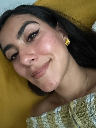 This is a close-up, low-angle selfie of a young woman with dark hair and light skin. She is lying on her side on a textured yellow surface, possibly a sofa or bed. Her head is tilted, and she is looking directly at the camera with a slight smile. She is wearing a summery, off-the-shoulder top with a pattern of green, white, and beige stripes. Her makeup includes winged eyeliner and a neutral-toned lipstick. A notable detail is a delicate gold earring in the shape of a starburst or flower. The lighting suggests it is daytime, possibly indoors, with soft illumination. The overall mood is relaxed and intimate. There is no visible text in the image.