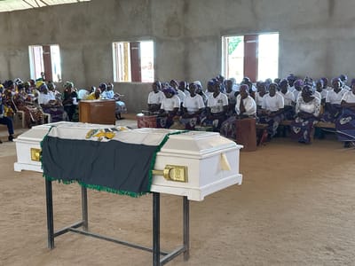 An indoor gathering is captured in this image, likely a funeral service, taking place on the grounds of a church or community hall in Kufana, Nigeria. A white coffin, draped with a dark green cloth adorned with a green fringe and a white cloth featuring a crest, rests on a metal stand in the foreground. The room is sparsely furnished with concrete walls and a corrugated metal roof. Natural light streams in through several windows, illuminating the space. A congregation of people, predominantly women dressed in traditional African attire and headscarves, are seated on benches, facing the coffin. Many are wearing white tops, suggesting a uniform for the occasion. Some individuals hold pamphlets or religious texts, engaged in prayer or song. The atmosphere appears somber and respectful. The ground is covered with sand.