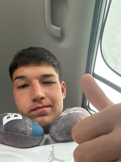 The image is a close-up, selfie-style shot taken from a low angle, possibly inside a vehicle. A young male with short, dark hair is looking at the camera, with his face filling most of the frame. He is giving a thumbs-up gesture with his right hand, which is positioned in the foreground. Two spherical objects, appearing to be plush toys designed to resemble American footballs, are visible on either side of the lower part of his face. The background suggests an indoor environment with a textured ceiling panel and a window with a sunshade on the right side, which has a faint, abstract pattern. The overall lighting is somewhat diffused, possibly from an overcast day or the interior lighting of the vehicle. The image does not provide any specific visual cues that would indicate a location within the city of Mirovo, Bulgaria.