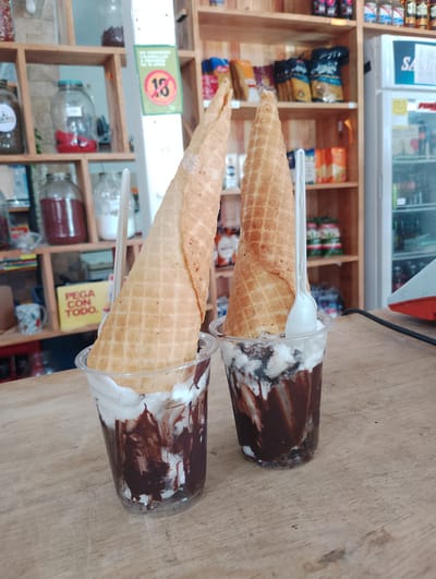 The image displays two ice cream desserts, each presented in a clear plastic cup filled with white ice cream and drizzled with chocolate syrup, topped with a large, inverted waffle cone. A white plastic spoon is inserted into each cup. The desserts rest on a worn, light brown wooden counter.
The background shows the interior of what appears to be a small shop or convenience store in El Espinal, Venezuela. Wooden shelves line the back wall, stocked with a variety of goods. On the left, several large glass jars are visible, containing different colored liquids or preserves. To the right, various packaged food items, including what looks like snacks and possibly pasta, are neatly arranged on the shelves. A prominent green sign with a red circle and the number "18" clearly states in Spanish: "NO VENDEMOS CIGARRILLOS A MENORES DE 18 AÑOS" (We do not sell cigarettes to minors under 18 years old). Below this, a yellow sign reads "PEGA CON TODO." (Sticks with everything.). On the far right, a white commercial refrigerator is visible, stocked with beverages, with the letters "SA" partially discernible on its side.
The setting suggests a casual, local business. No people are visible, and the activity is implied to be the preparation or sale of these ice cream treats. The lighting is even, suggesting an indoor, likely daytime, environment. The overall scene is static, focusing on the tempting desserts within the context of a bustling small shop.
