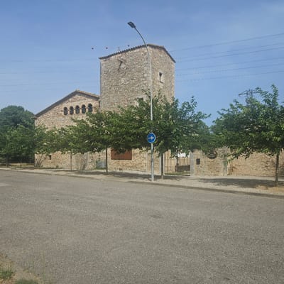 The image depicts a sunny day in what appears to be a rural or historical setting, likely in Girona, Spain. In the foreground, a paved road with a gravel shoulder runs horizontally across the frame. Beyond the road, a stone building with a tiled roof stands prominently. This building features a square tower on its right side and a lower section with arched windows on its left. Several leafy green trees are planted in a line in front of the building, casting dappled shadows on the ground. A modern street lamp with a curved arm stands near the tower.
To the right of the building, there is a metal gate and a stone wall. A circular blue traffic sign with a white arrow pointing to the right is mounted on the street lamp pole. On the wall of the building, there is a wooden sign with some text, possibly indicating a restaurant or establishment. In the sky above, there are power lines stretching horizontally and a few red spherical objects hanging from wires. The sky is clear and blue, suggesting good weather and likely mid-morning or afternoon. The overall scene conveys a sense of calm and history, with the modern elements like the street lamp and power lines juxtaposed against the ancient stone architecture.
