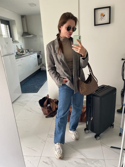 A person takes a mirror selfie indoors in Kranj, Slovenia. The selfie captures the person in a plaid blazer, brown shirt, jeans, and sneakers. They are wearing sunglasses and holding a phone with a green case. Next to them is a black suitcase and a brown handbag. The floor is white marble. Behind the person is a kitchen with white cabinets and a dark backsplash. A framed picture hangs on the wall above. A brown bag sits on the floor next to the person's feet.