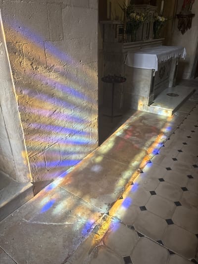 The image depicts the interior of a church in Maillane, France. Light streams from stained-glass windows, casting colored patterns on a stone wall and floor. An altar with white cloth and flower arrangements is visible in the background. The floor is tiled in a grid pattern of light and dark squares. A metal cross-shaped object is also near the altar. The wall has a rough, textured surface.