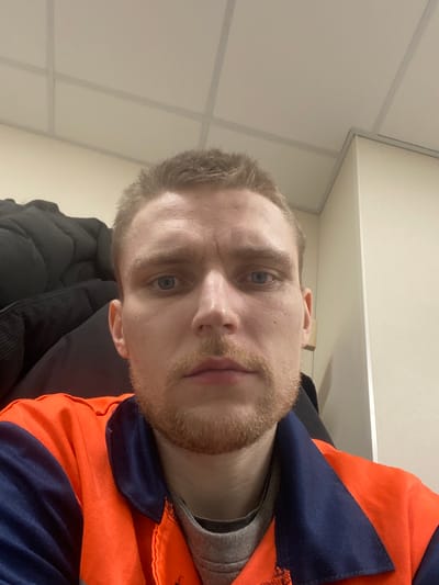 A male individual, appearing to be in his 20s or early 30s, is depicted in a close-up, head-and-shoulders portrait. He has short, light brown hair, blue eyes, and light stubble or a short beard. His expression is neutral, and he is looking directly forward. He wears an orange and navy blue two-tone work garment over a grey t-shirt. The background on the left features a dark, possibly black, puffy jacket or coat. On the right, a light-colored wall or cabinet with a vertical edge is visible. The overhead area reveals a light-colored grid-style suspended ceiling. The scene takes place indoors in the city of Nikol'skoe, Russia, likely within an office or a similar institutional setting.
