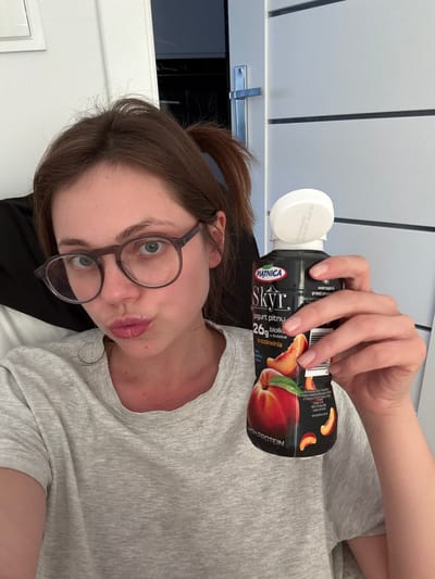The image shows a young woman lying down and holding a bottle of Piątnica Skyr yogurt with a peach flavor. She is wearing glasses and a grey t-shirt. Her hair is brown and pulled back. The background appears to be indoors, with white walls and what looks like a closet door. The lighting suggests it could be daytime. The bottle has text in Polish that says "Skyr jogurt pitny 26g białka w butelce brzoskwinia," which translates to "Skyr drinking yogurt 26g protein in a bottle peach." There is also text indicating "HIGH PROTEIN." The woman's expression is somewhat playful, with her lips puckered. The image orientation is sideways.
