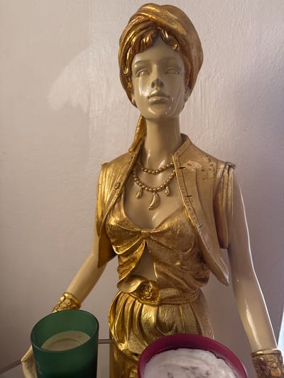 A golden statue of a woman in Middle Eastern attire stands against a white wall in Büyükçekmece, Turkey. The statue depicts the woman wearing a turban, a vest, and a flowing skirt, adorned with necklaces and bracelets. Her skin is a pale, glossy finish, contrasting with the metallic gold of her clothing and accessories. In the foreground, a green glass candle holder partially obscures the lower section of the statue, while a red candle holder is visible to the right. The lighting casts a soft shadow on the wall behind the statue.