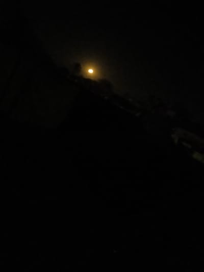 The image portrays a nighttime scene in Al-Mawasi, Palestinian Territories, characterized by extremely low light conditions. A single, prominent, circular light source, exhibiting a yellowish-orange hue, is visible in the upper-left quadrant, consistent with the moon or a distant artificial lamp. Faint, dark, amorphous silhouettes, possibly indicative of trees or architectural elements, are barely discernible directly below and to the right of this light source. The vast majority of the frame, particularly the lower and right portions, is undifferentiated black, signifying a profound absence of light and detail. No people, discernible actions, or specific objects beyond those mentioned are present.