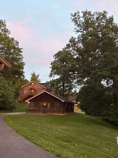 The image shows a scene at dusk in Washington Township, USA, featuring several wooden buildings nestled among lush green trees. A paved path curves through the foreground, leading towards a small, dark brown cabin with a screened-in porch. The cabin has interior lights on, casting a warm glow. Behind this cabin, a larger, multi-story wooden building with a prominent set of outdoor stairs is visible. This building also has lights on, suggesting occupancy. The sky above is a soft blend of pale blue and pink hues, indicative of twilight. The surrounding environment is predominantly foliage, with large trees dominating the right side of the frame and a dense growth of smaller trees and bushes on the left. The grass in the middle ground is short and dotted with small white flowers. The overall impression is of a tranquil, rustic setting, likely a retreat or lodging facility.