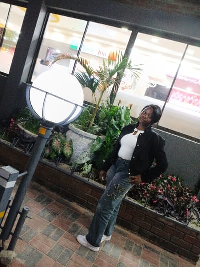 An adult woman is captured standing outdoors at night, posing casually beside a modern building. She is seen from the waist up, smiling faintly towards the camera. She wears a dark jacket over a white top, blue jeans with decorative stitching or embellishments on the thighs, and white sneakers. Her left hand is placed near her hip, and her right arm is slightly bent.

The setting appears to be a paved walkway or sidewalk next to a commercial building, possibly a shopping center or mall in Marondera, Zimbabwe. A large, spherical outdoor lamp with a grid pattern, emitting a bright white light, dominates the lower foreground. Behind the woman and the lamp are planters filled with various green foliage, including a small palm-like plant and broad-leafed plants, some with red or pink patterns.

The building behind the plants features large glass windows that reflect bright interior lights, indicating an evening or nighttime scene. Through the reflections, faint outlines of interior signage can be discerned. Part of a pink sign is visible in the upper right, possibly displaying the word "Creamy," and another fragmented sign below it may contain "Yummy Fun." The ground is paved with rectangular bricks in shades of brown and grey, and a low brick wall separates the planted area from the walkway. The overall atmosphere suggests a well-lit, public commercial area during off-peak hours.