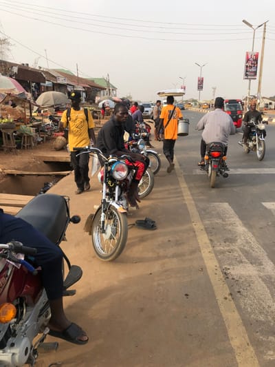 A bustling street scene shows multiple motorcycles parked or moving on a road. Several people are visible; some are on the motorcycles, while one carries containers on their head. Street vendors and shops line the side of the road, suggesting a market area. Overhead wires and billboards add to the chaotic yet vibrant urban environment. The photo is taken from a slightly elevated position, possibly from another vehicle or building.