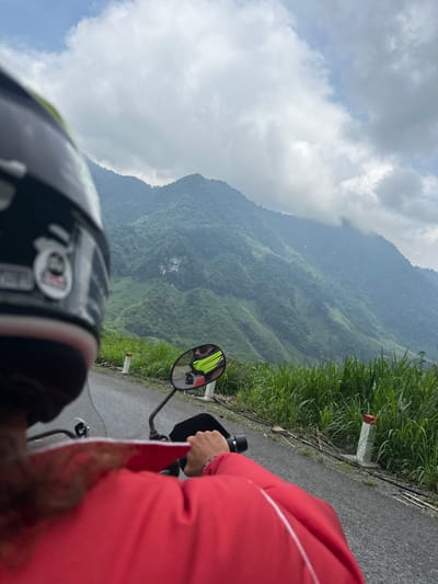 The image captures a first-person perspective from a motorbike, showcasing a scenic ride through a mountainous landscape in Minh Sơn Commune, Vietnam.
In the foreground, the back of a black and white motorcycle helmet is visible on the left, featuring a sticker that resembles the "AGV" logo. To the right, the arm and torso of a person wearing a bright red jacket are seen, likely a passenger. The motorbike's handlebars and a rearview mirror are also prominent. The mirror reflects another individual, presumably the driver, wearing a bright yellow or lime green reflective vest.
The motorbike is on an asphalt road that curves alongside the mountainous terrain. Lush green grass and dense foliage line the roadside, and a concrete road marker with a distinctive red and white painted top is visible. Beyond the road, vast, verdant mountains covered in thick forests stretch into the distance, partially obscured by mist or clouds, indicating a high altitude and potentially humid environment. The sky is partly cloudy with some clear blue patches, suggesting it is daytime. The scene conveys a sense of adventure and travel through a beautiful natural setting.