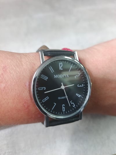 A close-up shot shows a black analog wristwatch with a silver bezel on a person's wrist. The watch face displays white Arabic numerals and hour markers, with the brand name "MODIYA" and "QUARTZ" printed below the 12 o'clock position. The hour hand is pointing slightly past the 12, and the minute hand is pointing at the 12. The watch has a black leather strap. The background is a light-colored, textured surface, possibly a wall or countertop. There are no identifiable landmarks or context clues to suggest the location is in Mikhnevo, Russia.
