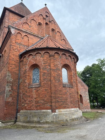 This is a close-up, exterior, eye-level, full-shot photograph of a section of a red brick church in Bassum, Germany. The church features a rounded apse with two arched windows that appear to be made of frosted glass with a diamond pattern. The brickwork is detailed, with decorative arched brick patterns above the windows. The roof is tiled in a terracotta color. A grey stone base or foundation is visible at the bottom of the apse. To the right of the church, a large, mature green tree is prominent, and the sky is overcast and grey. The foreground shows a grey asphalt or gravel path. The image captures architectural details of the historic building.