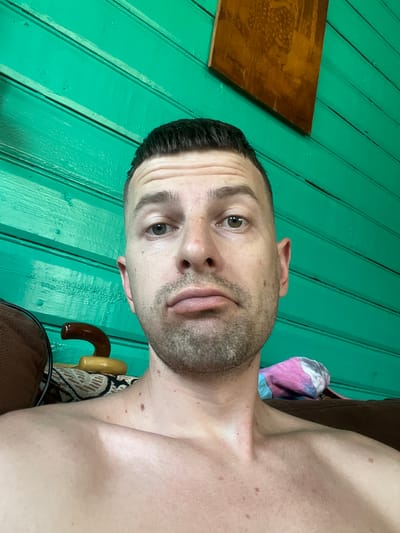 A man with short, dark hair and a beard is taking a selfie in front of a teal wooden wall. He is shirtless and looking at the camera with a neutral expression. Behind him, to his left, a brown chair is visible, with a patterned fabric draped over it and a dark wooden handle protruding. To his right, a pink, blue, and white tie-dyed fabric is draped. A small, framed piece of artwork hangs on the wall above and to the right of his head. The man's skin is fair with some freckles and moles. He has light brown eyes.
