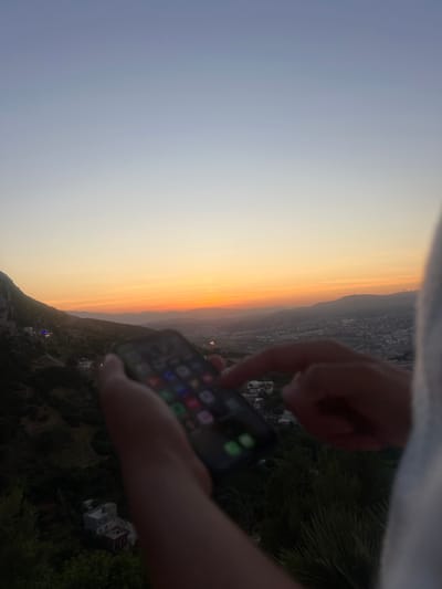 The image depicts a person holding a smartphone, with a vibrant sunset over a mountainous city as the backdrop. In the foreground, a pair of hands grasps a black smartphone, its screen illuminated with a grid of colorful application icons. A thumb is poised over the screen, suggesting interaction. Faint green foliage is visible near the bottom left, partially obscured by the hand.

The background unfolds a picturesque view during what appears to be late dusk or sunset. To the left, dark, tree-covered hills rise, dotted with a few indistinct buildings. The horizon is dominated by a sweeping gradient of warm colors, from bright orange and yellow directly above the distant mountains to softer purples and blues higher in the sky. These mountains extend across the mid-ground, their forms silhouetted against the brilliant light. Below the mountain range and extending towards the foreground, a city spreads out, its urban sprawl visible in the dimming light. The sky is clear, suggesting fair weather. The scene conveys a moment of quiet observation, blending human technology with the grandeur of nature. The location is specified as Tétouan, Morocco. There is no legible text visible on the phone screen or elsewhere.