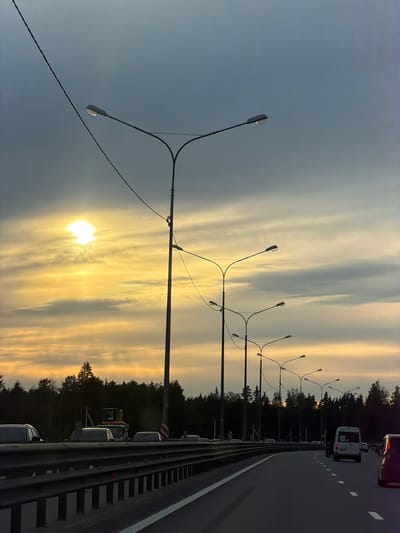 The image depicts a highway in Gory, Russia, at sunset. The scene is characterized by a dramatic sky filled with soft, golden clouds and the bright, low sun. Several modern streetlights with multiple arms line the side of the road, silhouetted against the colorful sky. The highway itself is paved, with clear lane markings. A metal guardrail separates the road from the adjacent forested area.
Several vehicles are visible on the highway, including a white minivan in the distance and other cars further down the road. The dense line of trees bordering the highway suggests a rural or suburban setting. The overall mood is serene and contemplative, highlighted by the warm glow of the setting sun. There is no visible text in the image.