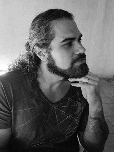 A black and white medium shot features a single adult male, positioned off-center left and looking towards the right side of the frame. He has long, dark, curly hair with visible grey streaks, tied back into a bun, and a full, dark beard and mustache. His right hand, adorned with a plain band ring on the fourth finger, is raised with fingers touching his chin. He wears a dark V-neck t-shirt featuring a light-colored geometric pattern of intersecting lines. The background consists of a plain, light-colored, textured wall, with a darker, textured surface visible at the bottom right. The scene is located in Villa San Andrés, Argentina.