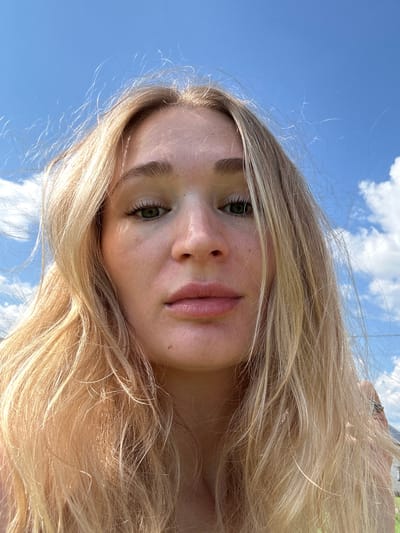 A young woman with blonde hair and green eyes looks directly at the camera, her face filling most of the frame. She has fair skin with freckles and appears to be outdoors, with a bright blue sky and some white clouds visible behind her. Her hair is slightly windblown, framing her face. The lighting suggests it is daytime, and the overall scene is casual and natural. There are no identifiable landmarks that would suggest a specific location within the city of Mokino, Russia.