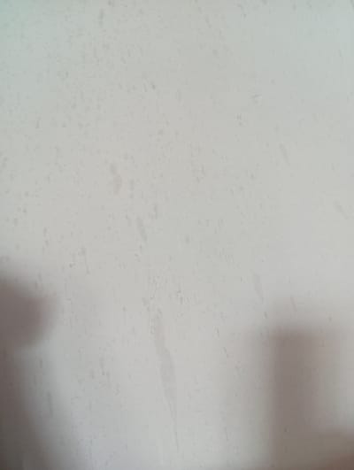 The image displays a close-up, overhead view of a wall with a light-colored, textured surface. There are numerous small, irregular dark spots scattered across the wall, resembling splashes or stains. Faint, lighter streaks and lines are also visible, suggesting wear or perhaps a faint pattern. In the lower left and right portions of the image, soft, diffused shadows are present, indicating the presence of objects outside the frame that are casting shadows onto the wall. The lighting appears to be ambient and diffused. The image is taken indoors, likely in a building in Romanovka, Kyrgyzstan, though no specific landmarks or context are discernible. The wall itself appears to be made of concrete or a similar material. No people, animals, or specific objects are clearly identifiable within the frame.