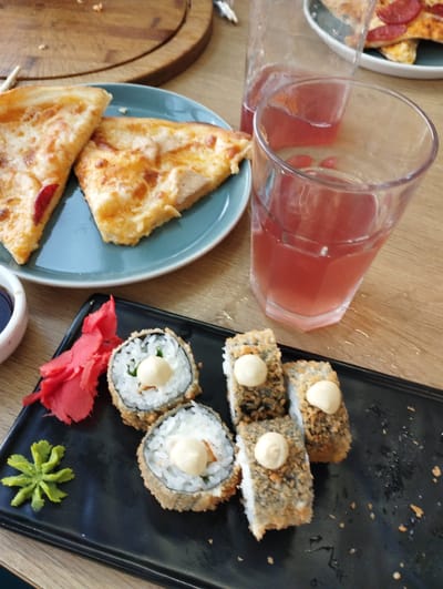 An overhead, medium shot captures a wooden table laden with food and drinks. In the foreground, a black rectangular plate holds several pieces of sushi, each topped with a dollop of white sauce. To the left of the sushi, pickled ginger, colored a vibrant pink, is artfully arranged, and a small mound of green wasabi sits nearby. Behind the sushi, a glass of pink beverage with ice cubes sits on the table, next to another glass containing a similar drink. A blue plate in the background contains two slices of pizza with melted cheese and pepperoni. A wooden cutting board, partially visible at the top of the frame, suggests additional food items may have been present. The scene is set in a dining establishment, likely a restaurant, in Talas City, Kyrgyzstan, as indicated by the provided location context. The lighting is bright, and the focus is sharp, highlighting the textures and colors of the food. There are no people visible in the image.