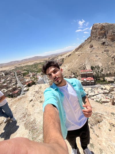 A young man takes a selfie overlooking the city of Ma'loula, Syria. He stands on a rocky, cliffside perch. He wears a white t-shirt, an open light blue button-down shirt, and dark pants. The landscape stretches into the distance with buildings and roads visible below. The sky is clear and blue. Part of a rocky cliff face is visible to the upper left. The city is situated in a valley.