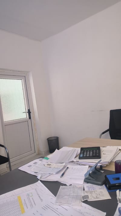The image depicts an interior scene, likely an office, with a desk in the foreground covered in papers, a calculator, a hole punch, and a pen. A black office chair is partially visible to the right. To the left, a white door with a frosted glass panel and a black handle is present. A black mesh trash can stands near the door. The walls and ceiling are painted white. The scene appears to be a disorganized workspace, suggesting recent activity or ongoing tasks. The provided context indicates this location is in the city of Булак-Башы, Kyrgyzstan.