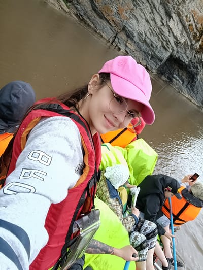In Idrisovo, Russia, a group of people are on a boat with a rocky cliff face in the background. The water appears murky brown. A young woman in the foreground is taking a selfie. She is wearing a pink baseball cap, glasses, and a red life vest over a gray sweatshirt with black lettering. Other individuals are visible in the boat, some wearing life vests and head coverings, and one person is holding a smartphone. Bright yellow and green life vests are stacked in the boat.