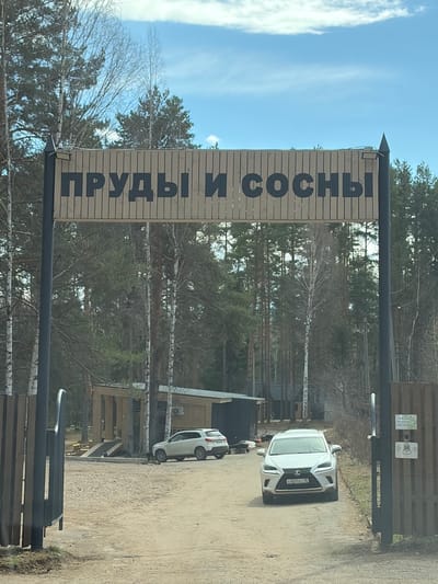 This media file captures an outdoor scene at the entrance of a facility named "ПРУДЫ И СОСНЫ" (PRUDY I SOSNY), meaning "Ponds and Pines," located in Mishkino, Russia. The image is taken during the daytime under a bright, clear blue sky with some wispy clouds, indicating fair weather.

A large, wooden sign is centrally positioned, displaying the name "ПРУДЫ И СОСНЫ" in prominent dark, capitalized letters on a light wooden background. This sign spans above a dark-colored wooden gate or fence structure, which serves as the entrance to the property. On one of the gate posts, small signs are affixed; one has a speaker icon, and another green sign partially shows text that appears to read "ВЫЗОВ ОХРАНЫ" (CALL SECURITY) and "ПРИЯТНОГО ОТДЫХА" (ENJOY YOUR STAY).

Two white vehicles are visible on a dirt or gravel road leading into the entrance. A white Lexus SUV is closer to the gate, displaying a Russian license plate with the registration "C601EC 18". Another smaller white car is further in the background. The surroundings are heavily wooded, dominated by numerous trees, including birches with their distinctive white bark and various evergreen conifers, suggesting a natural, forest-like setting. Scattered wooden structures, possibly cabins or recreational buildings, are discernible among the trees. There are no people visible, and the scene depicts a static view of the entrance area.