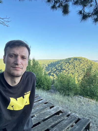 A man is taking a selfie on a wooden structure outdoors with a landscape of trees in the background. The man is wearing a black t-shirt with a yellow logo. The landscape consists of rolling hills covered in green trees under a clear blue sky. There are pine branches visible in the upper corners of the image. The wooden structure appears to be made of pallets. The location is the city of Pryvorottia Druhe, Ukraine.