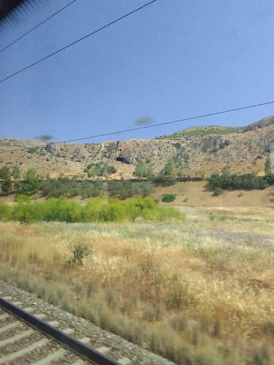 This media file depicts a rural landscape under a clear blue sky, suggesting daytime. The scene is viewed from a moving train, indicated by the blurred foreground of railway tracks. In the midground, a strip of vibrant green bushes stretches horizontally, contrasting with the dry, golden grasses and vegetation behind them. Beyond this, a hilly terrain rises, characterized by rocky outcrops and sparse vegetation. A notable feature on the hillside is a dark opening that appears to be a cave entrance. The overall setting suggests a Mediterranean climate, with the dry vegetation hinting at a warm, possibly summer season. There is no visible text in the image. The image captures a serene, natural environment, likely in the Municipal Unit of Fili, Greece.