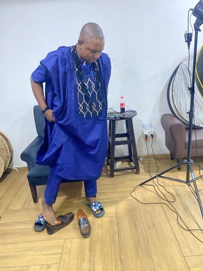 A man in a blue traditional African outfit stands on a wooden floor. He wears casual sandals, with a pair of dress shoes beside his feet. A black stool stands behind him, with a Coca-Cola bottle and a cell phone placed on top. A lighting rig is positioned in the background, along with a brown leather couch. A woven basket is partially visible in the lower left corner. The wall is white with a visible outlet.