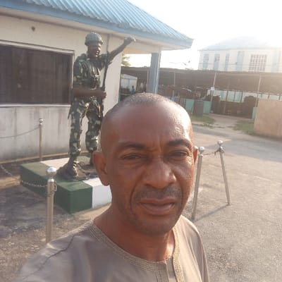 In Koko, Nigeria, a dark-skinned adult male with a shaved head, wearing a light brown traditional garment, is depicted in a selfie. He stands outdoors on a paved surface under bright sunlight. Behind him, to the left, is a life-sized statue of a soldier in camouflage uniform, helmet, and boots, holding a long implement diagonally with his right arm raised. The statue is positioned on a two-tiered pedestal (dark green base, white top) and partially enclosed by a low barrier of silver posts connected by chains. Immediately behind the statue is a single-story building with a light-colored facade, a light blue corrugated metal roof, and a dark-tinted window. To the right of the statue, an open area contains multiple dark cylindrical objects, possibly barrels, situated behind a mesh or slatted fence. Further in the background, a light-colored, multi-story building with numerous windows is visible against a bright sky.