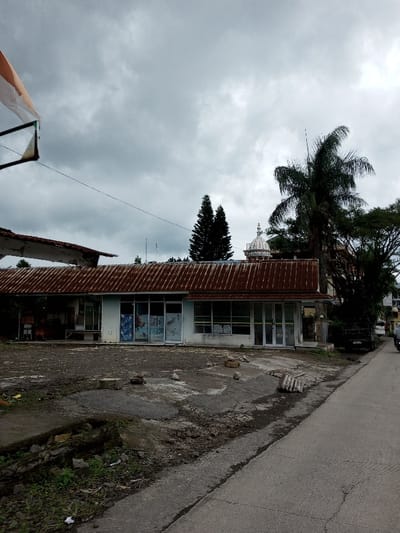 The image depicts an exterior street view in Melakudikadu, India, under an overcast sky with grey clouds. The foreground features an uneven, cracked paved and dirt lot with scattered rocks and debris, including a partially visible corrugated metal sheet. A small, overgrown drainage ditch runs along the bottom-left. To the right, a paved road extends diagonally into the background.
The central subject is a single-story building with a light-colored facade and a prominent, rusty, corrugated metal roof. The building features multiple large glass windows and doors across its front, some containing visible posters or promotional materials. On the far left, an older, partially deconstructed or damaged roof section extends, revealing an underlying structure.
In the mid-ground, a dark-colored utility truck is partially visible on the right side of the road, obscured by trees. Further down the road, a person in a light-colored top is partially visible near a motorbike or scooter. Overhead, thin wires traverse horizontally.
The background includes dense green foliage from various trees, notably a tall, dark green coniferous tree and a large palm tree on the right side of the frame. A white domed structure with a finial is visible above the building, positioned between the trees. In the top-left, a portion of a white and orange flag or banner is visible, attached to an unseen pole structure, with a triangular frame hanging below it. No explicit interactions are discernible among the visible subjects.
