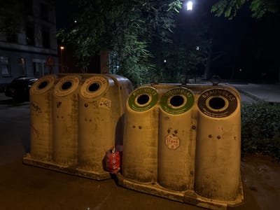 The image shows a nighttime outdoor scene in Duisburg, Germany, featuring two large, cylindrical glass recycling bins. The bin on the left is labeled "WEISSGLAS" (white glass) and has three circular openings. The bin on the right is divided into two sections: "GRÜNGLAS" (green glass) and "BRAUNGLAS" (brown glass), each with a circular opening. Both bins are weathered and show signs of graffiti. A red and white patterned reusable shopping bag is placed at the base of the left bin. In the background, there is a building with illuminated windows, trees, a streetlight, and a street sign with a pictogram of children walking. The ground appears to be asphalt or concrete, and a parked car is partially visible on the left side of the image.