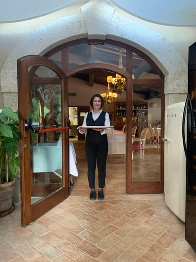 A woman in a black vest and black pants, likely a server, stands in a doorway holding a silver tray with a white cloth. She is looking towards the camera with a neutral expression. She is inside what appears to be a restaurant or establishment with a rustic aesthetic. The doorway is framed by an arched stone structure and wooden doors. Through the glass of the doors, a dining area with tables and chairs is visible. In the foreground to the left is a large potted plant, and to the right is a cream-colored Smeg refrigerator. The floor is paved with terracotta tiles. The overall impression is that of a charming establishment in a historic Italian town, possibly Castiglione d'Orcia given the context.