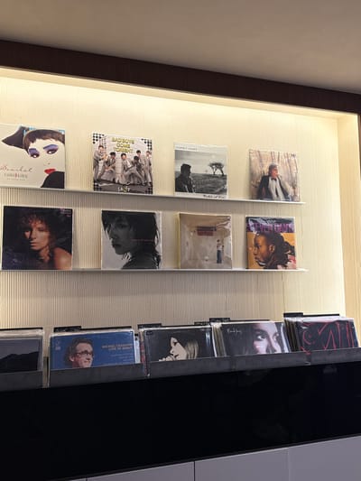 The image depicts a retail display of vinyl records within what appears to be a store or establishment. The display consists of several shelves, each holding multiple vinyl record albums. The albums are arranged in a horizontal manner, with their covers facing outward. The lighting suggests an indoor environment, possibly with track lighting or ambient illumination from above. The background wall has a textured, vertical ribbed pattern.
The upper shelves feature albums with distinct artwork and titles. Notable albums visible include:
- An album with a close-up of a woman's face with bold makeup. Text below reads "Scarlet" and "NAOKO KAWAI".
- An album titled "BAD BOYS COME TONIGHT", featuring a group of five men in matching suits.
- An album with the title "COREY HART" and "Fields of Fire".
- An album featuring a man in formal attire.
- An album with a black and white portrait of a woman, titled "Barbra Streisand Wet".
- An album with a black and white portrait of a woman, titled "Sakura, Paradise Callin'".
- An album with an image of a person standing in a sparsely furnished room.
- An album with a portrait of a person's head in profile, titled "STEVEN DANS".
The lower shelves display more albums, some of which have visible titles:
- An album titled "MICHEL LEGRAND LIVE IN JAPAN".
- An album with the title "Nordy Jays".
- An album with a prominent red graphic design.
The overall scene is orderly and organized, typical of a commercial setting for music retail. There is no clear indication of the specific city or district within China, other than the query prompt mentioning Jing'an District.
