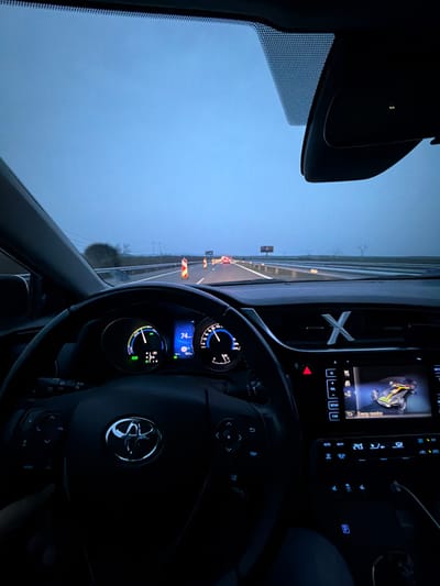 The scene depicts the interior of a vehicle and the highway ahead, identified as being in Krustina, Bulgaria, during what appears to be either dusk or dawn under an overcast sky.

**Vehicle Interior:** The perspective is from the driver's or front passenger's seat. A dark, multi-function Toyota steering wheel with an illuminated emblem is central in the foreground. The driver's left hand is visible on the lower left portion of the steering wheel. The illuminated instrument cluster behind the steering wheel displays a speedometer indicating approximately 74 units (likely km/h) and a power meter showing current energy usage. An infotainment screen on the center console displays an "Energy monitor" graphic of a hybrid car with active power flow. Below this screen are climate controls showing "24.5" (likely Celsius) and other buttons. An illuminated red hazard light button is visible. A metallic "X" shaped object rests on the dashboard above the center console. The base of an overhead rearview mirror is visible at the top center of the windshield.

**Exterior Environment:** Through the windshield, a multi-lane highway extends into the distance. The sky is a uniform pale blue-grey. The road ahead features multiple orange and white striped traffic cones, indicating a designated work zone or lane modification. Distant red taillights of other vehicles are visible. On the left side of the highway, a roadside area with sparse vegetation and what appear to be power lines or utility poles is visible. On the right, a guardrail parallels the road, and two billboards are visible in the far distance. The highway appears to have at least two lanes in the direction of travel, separated by a median.