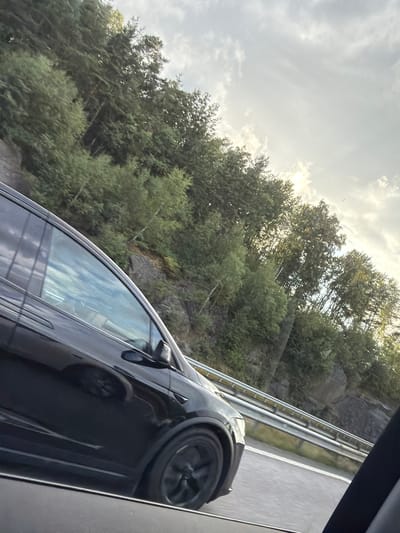 The image, taken from inside a vehicle on a sunny day, shows a black Tesla SUV driving on a highway. The car is in motion, with its wheels blurred. The highway has a metal guardrail and white lane markings. To the right of the highway, a rocky hillside covered in dense green trees rises steeply. The sky is partly cloudy, with sunlight breaking through the clouds, casting a bright glow. The overall scene suggests travel through a scenic, possibly mountainous or hilly, natural environment. The location is identified as Åsa, Sweden.