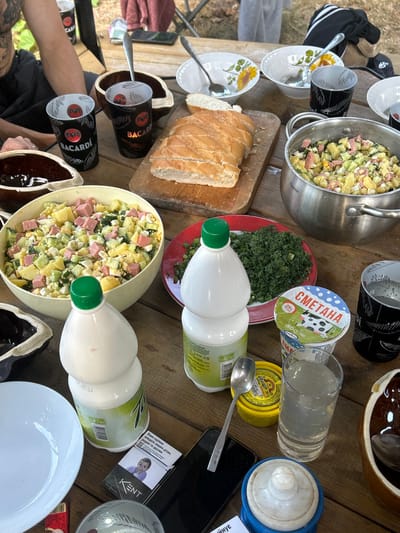 The image captures an outdoor meal scene on a weathered wooden plank table in the city of Dibrova, Ukraine. The table is set with multiple dishes and beverages.
Two large containers, one a pale yellow bowl and the other a metallic pot, are filled with a diced salad consisting of potatoes, ham or sausage, and cucumbers, consistent with Olivier salad. A sliced loaf of light-colored bread rests on a wooden cutting board. Two red-rimmed white plates contain finely chopped green onions and dill. A white plastic container of "СМЕТАНА" (sour cream) labeled "21%" is present. A yellow round container, possibly for a condiment, has a metal spoon resting on it.
Beverages include two white plastic bottles with green caps, one labeled "1.0 L." Several black Bacardi-branded cups are visible, along with a clear glass containing a light-colored liquid. Multiple white ceramic bowls, some adorned with sunflower patterns, and dark brown serving dishes are also on the table. Metal spoons are scattered among the items.
Personal effects include a black smartphone, a pack of "KENT" cigarettes displaying a health warning with an image of a child, and a red lighter. A blue and white object, possibly an ashtray or small container, is also present.
Partial views of two individuals are visible: an arm and tattooed torso of a person in a dark top on the left, and an arm and leg of another person in light clothing on the right. The background shows blurred green foliage, confirming an outdoor setting under natural light.