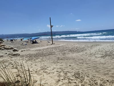 The image shows a beach scene in Kiryat Yam, Israel, on a clear, sunny day with a bright blue sky and scattered white clouds. The foreground is dominated by a sandy beach with ripples and footprints. Some dry, thin vegetation is visible in the lower left corner.

In the mid-ground, several blue beach umbrellas provide shade for people sitting on the sand. Some beach chairs and towels are also visible. A tall wooden pole with a light fixture stands vertically in the middle of the beach. Further back, the ocean waves crash onto the shore, and several people are visible in the water, some swimming or wading.

The background features a distant city skyline along a hilly coastline, with buildings and what appear to be industrial structures, possibly cranes at a port. The sea stretches to the horizon, meeting the sky under a clear blue expanse.