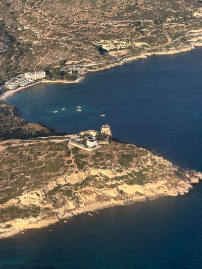 This aerial view captures a coastal landscape near Cagliari, Italy. The scene is dominated by the deep blue of the sea meeting a rocky, arid coastline dotted with sparse vegetation. In the foreground, a rocky promontory juts into the sea, crowned by a white and beige structure that appears to be a lighthouse or a watchtower, with a few smaller buildings clustered around its base. A road winds along the coastline, with several parked cars visible near the tower. Further along the coast, a bay shelters a small beach and a cluster of low-rise buildings, likely hotels or residences, characterized by their light-colored facades. Several small boats are anchored in the calm waters of the bay. The hilly terrain behind the coastal developments is covered in dry, scrubby vegetation and crisscrossed by dirt paths.