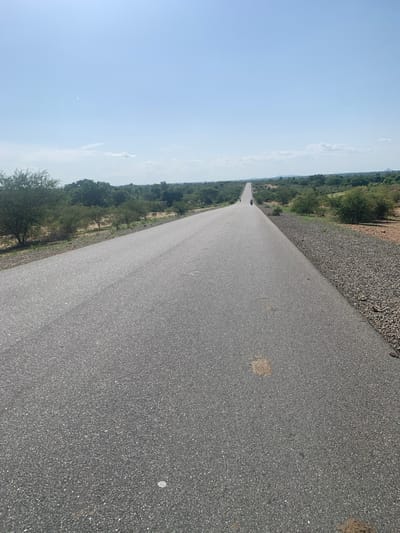 The image shows a straight, paved road stretching into the distance. The road is dark asphalt and appears to be in good condition. On either side of the road, there is dry, scrubby vegetation and scattered trees, suggesting a rural or semi-arid environment. To the left, a vast expanse of light blue sky dominates the frame, with a few wispy clouds visible. To the right of the road, there is a narrow strip of gravel or small stones.
In the distance, on the road, two figures on motorcycles are visible, heading away from the viewer. This indicates that the scene is likely taking place during daylight hours. The overall impression is one of a journey or travel through a somewhat desolate, yet open, landscape. The lack of prominent landmarks makes it difficult to pinpoint an exact location, but the environment is consistent with parts of Nigeria. There is no visible text in the image.