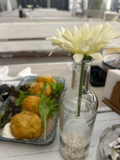 The image depicts a close-up view of a table setting, likely in a cafe or restaurant in Aytos, Bulgaria. The primary subjects are a single white flower in a clear glass bottle, a plate of food, and an ashtray with cigarette butts.

The flower, appearing to be a chrysanthemum, is pale yellow and is the focal point, placed in a repurposed glass bottle that also contains small white pebbles. The bottle itself is lying on its side, with the flower stem extending from the opening.

In front of the bottle, a rectangular plate holds several golden-brown, round food items, possibly croquettes or falafel, garnished with fresh green lettuce. A small dollop of white sauce or dip is visible on the plate.

In the background, an ashtray with several smoked cigarette butts is partially visible. The overall scene suggests a casual setting, possibly a break or a meal. The lighting appears to be natural, implying it could be daytime. The presence of the ashtray might indicate an outdoor or smoking-permitted area. No specific text is clearly legible in the image, although there might be faint branding on the glass bottle. The camera angle is from above, looking down at the table.