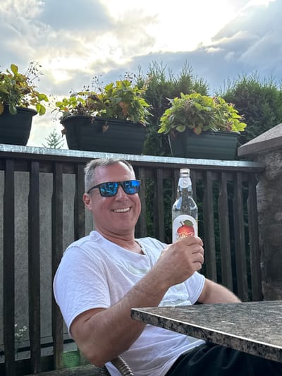A man is holding a clear glass bottle with a light-colored liquid and a red apple label. He is wearing a white t-shirt and sunglasses with blue lenses. The man is smiling and looking at the camera. He is sitting at a table outdoors. In the background, there are potted plants and a wooden fence. The sky is cloudy. The photo was taken in Rūdiškės, Lithuania.