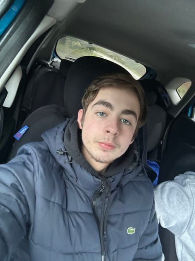 A close-up selfie-style image depicts a young male seated in the front passenger seat of a vehicle. The individual, appearing to be in his late teens or early twenties, has light skin, light brown hair, prominent eyebrows, and green or blue-green eyes. He has minimal facial hair, consisting of light stubble on his chin and upper lip. He is wearing a dark blue hooded puffer jacket with a visible green crocodile logo (Lacoste) on the left chest, over a black hooded sweatshirt. His gaze is directed forward, presumably at the camera.
To the right of the primary subject, a portion of a second individual is visible, characterized by a grey hooded garment covering the shoulder and upper arm, suggesting another occupant in the vehicle.
The interior of the vehicle is visible, featuring dark-colored upholstery on the seats. On the headrest of the seat directly behind the primary subject, a blue tag with "Sparco" and "Corsa" branding is attached. Behind this seat, dark-colored luggage or bags are discernible in the rear passenger area.
Through the rear window of the vehicle, a blurred outdoor environment is visible, consistent with a natural or rural landscape featuring green foliage and distant trees under what appears to be an overcast sky. The specific city of Ribeira de Pena, Portugal, is not visually discernible within the image.