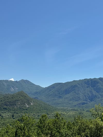 The image depicts a mountainside landscape under a clear, blue sky. Lush, green vegetation covers the slopes of the mountains, with trees visible on the right. The scene is likely located in a rural area. No people are visible. The location is in Sotonići, Montenegro.